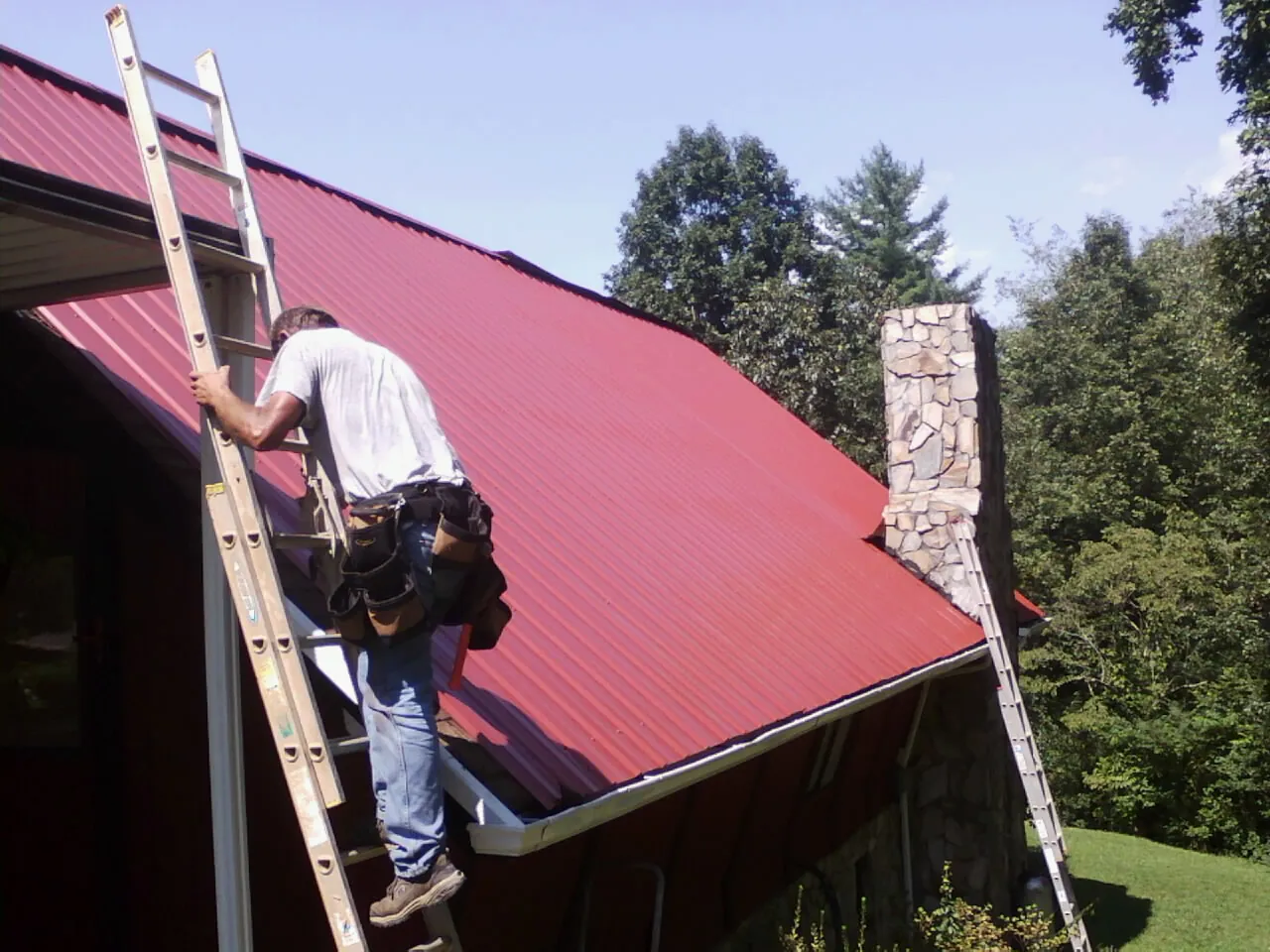 Roofer on a ladder beside a red metal roof and stone chimney — Soffit & Fascia Repair by Roofing Brothers in Mount Hope